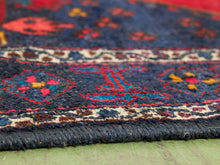 Load image into Gallery viewer, Close-up of a patterned rug corner showing a dark navy blue field with red, beige, yellow, and brown motifs, plus a fringed edge resting on a green surface.