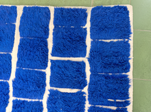 Load image into Gallery viewer, A close-up of a blue shaggy rectangular rug with a white border, laid on a green tiled floor.