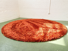 Load image into Gallery viewer, A large round shag rug in a warm rust-orange color, displayed on a green floor against a white brick wall.