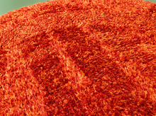 Load image into Gallery viewer, A close-up of a bright orange-red shaggy rug or carpet texture filling the frame, with a small portion of a pale green surface visible in the top-left corner.