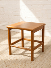 Load image into Gallery viewer, A square wooden stool with a warm medium-brown finish, featuring a slanted seat and front support bars, set on a textured beige rug against a white brick wall with soft sunlight and shadows.