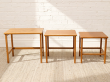 Load image into Gallery viewer, Three wooden nesting tables of varying heights in a warm light oak finish, arranged against a white brick wall on a textured beige rug.