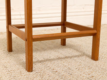 Load image into Gallery viewer, Close-up of a wooden stool or table leg frame with a warm medium-brown finish, standing on a woven beige rug against a white wall.