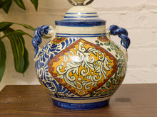 Load image into Gallery viewer, A hand-painted ceramic lidded jar with two handles, featuring a vibrant yellow central diamond pattern surrounded by blue, green, and white scrollwork, set on a wooden surface against a white brick wall.