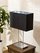 Load image into Gallery viewer, A modern table lamp with a matte black rectangular shade and a polished silver metal base, shown on a wooden side table against a white brick wall with a potted plant nearby.