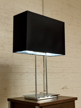 Load image into Gallery viewer, A modern table lamp with a large black rectangular shade, a polished silver metal base, and two slim vertical silver stems, shown resting on a wooden side table against a white brick wall.