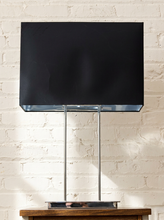 Load image into Gallery viewer, A modern table lamp with a large black rectangular shade supported by two slim vertical chrome legs, standing on a small metallic base against a white painted brick wall.