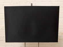 Load image into Gallery viewer, A large black rectangular canvas or panel displayed on a metal stand against a white painted brick wall.