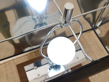 Load image into Gallery viewer, A close-up of a modern table lamp with a polished chrome metal frame and a glowing white globe shade, set on a glass-topped table.