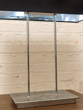 Load image into Gallery viewer, A modern table lamp with a rectangular black shade, two slim vertical chrome metal poles, and a square brushed-metal base, set against a white brick wall.