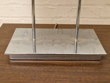 Load image into Gallery viewer, A close-up of a modern table lamp base featuring a square polished chrome metal platform and two vertical chrome rods, set on a dark wood tabletop against a light brick wall.