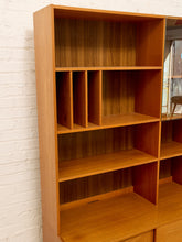 Load image into Gallery viewer, A mid-century modern wooden bookcase with warm orange-brown teak wood grain, featuring open shelves and a mirrored cabinet door on the right, set against a white painted brick wall.
