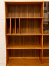 Load image into Gallery viewer, A mid-century modern wooden bookcase with warm honey-toned wood grain, featuring open shelves, vertical dividers, and a glass-front cabinet section on the right, set against a white brick wall.