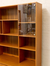 Load image into Gallery viewer, A mid-century modern wooden bookcase with a warm honey finish, featuring open cubby shelves on the left and a glass-front cabinet on the right with two small white knobs, set against a white brick wall.