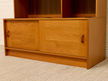 Load image into Gallery viewer, A mid-century modern wooden credenza or sideboard cabinet with two sliding doors and small round handles, shown in a warm honey-oak finish against a white brick wall and beige carpet.
