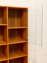 Load image into Gallery viewer, A wooden bookshelf with open cubbies and a clear glass door hinged on the right side, shown against a white brick wall.