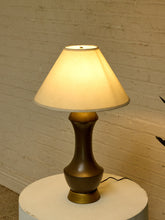 Load image into Gallery viewer, A table lamp with a warm white fabric drum shade and a dark bronze-colored ceramic base, illuminated against a white brick wall background.