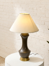 Load image into Gallery viewer, A table lamp with a warm white conical shade, a dark brown sculptural ceramic base, and a brass-colored bottom ring, shown lit on a white round pedestal against a white brick wall.