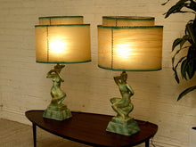 Load image into Gallery viewer, Two matching table lamps with sculptural green bases and beige drum shades, shown on a dark wood table against a white brick wall with a plant at the side.