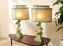 Load image into Gallery viewer, Two matching table lamps with sculptural green patina bases and beige drum shades with teal trim, displayed on a dark wood table against a white brick wall.