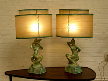 Load image into Gallery viewer, A pair of vintage-style table lamps with sculptural green female figure bases and beige drum shades with green trim, shown on a dark wooden table against a white brick wall.