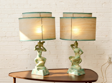 Load image into Gallery viewer, A pair of table lamps with sculptural green bases and beige drum shades with teal trim, displayed on a wooden table against a white brick wall.