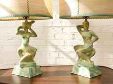 Load image into Gallery viewer, A pair of table lamps with sculptural green-and-bronze female figure bases, shown on a wooden surface against a white brick wall. Each lamp has a beige lampshade with a teal scalloped trim.