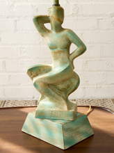 Load image into Gallery viewer, A sculptural table lamp featuring a stylized seated female figure in a distressed turquoise and cream finish, placed on a wooden surface against a white brick wall.