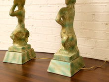 Load image into Gallery viewer, A pair of sculptural table lamps shaped like stylized female figures with a distressed turquoise and green patina finish, standing on square bases on a dark wood table against a white brick wall.