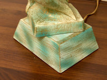 Load image into Gallery viewer, A close-up of a decorative lamp base made from stacked rectangular wood blocks with a distressed turquoise and beige finish, placed on a brown wooden tabletop. A matching distressed fabric-covered top piece rests on the blocks, and a tan cord extends from the upper right.