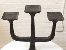 Load image into Gallery viewer, A black cast iron three-arm candelabra with square candle cups, shown on a white surface against a white brick wall background.