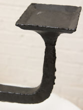 Load image into Gallery viewer, A close-up of a rustic black metal wall hook with a square-shaped top and a thick, textured stem, set against a light off-white brick wall background.