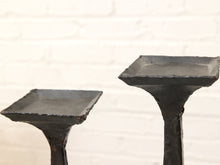 Load image into Gallery viewer, Two rustic black cast iron candlesticks with square tops, shown against a white brick wall background.