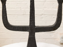 Load image into Gallery viewer, A close-up of a black cast iron trident candle holder with a rustic, textured finish, set against a white brick wall background.