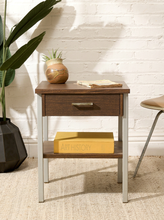 Load image into Gallery viewer, A mid-century modern two-tone nightstand with a dark brown wood top and drawer front, light gray metal legs, and a lower shelf. The nightstand is styled with a ceramic pot, a book, and a yellow box on a beige woven rug in front of a white brick wall.