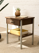 Load image into Gallery viewer, A mid-century modern two-tier nightstand with dark brown wood grain and silver metal legs, styled with a small potted succulent and a yellow book against a white brick wall background.
