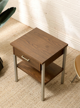 Load image into Gallery viewer, A two-tier wooden end table with a single drawer and a metal pull handle, set on a textured beige rug against a white brick wall.
