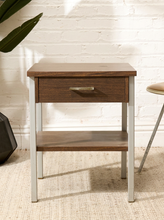 Load image into Gallery viewer, A two-tier wooden nightstand with a single drawer and a metal pull handle, set against a white brick wall background.