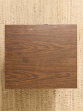 Load image into Gallery viewer, A square wood-look sample tile with a medium-to-dark brown grain pattern, displayed flat on a textured beige woven rug.