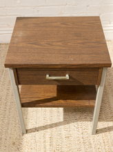 Load image into Gallery viewer, A mid-century modern wooden nightstand with a dark brown wood grain finish, featuring a single drawer with a silver metal pull handle and an open lower shelf, supported by slim silver metal legs.