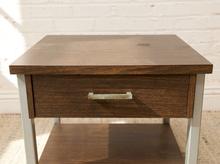 Load image into Gallery viewer, A mid-century modern wooden nightstand with a dark brown wood finish, a single drawer with a brass pull handle, and an open lower shelf on light gray metal legs, set against a white brick wall.