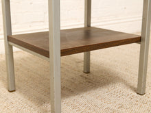 Load image into Gallery viewer, A close-up of a two-tier side table featuring a dark brown wood grain shelf and a distressed light gray metal frame, set on a beige woven rug against a white brick wall.