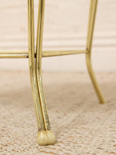 Load image into Gallery viewer, Close-up of a brass or gold-finished metal stand or table leg with a round beige foot, standing on a woven beige rug against a light neutral background.