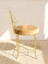 Load image into Gallery viewer, A gold metal barstool with a round tufted beige velvet cushion, set against a white brick wall on a textured beige rug.