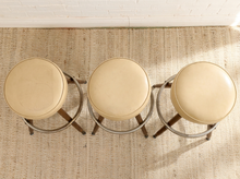 Load image into Gallery viewer, Set of three vintage bar stools with round cream upholstered seats, four splayed wood legs, and circular metal footrests