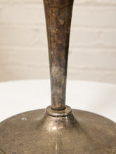 Load image into Gallery viewer, A close-up of a vintage metal table base or lamp stand with a wide circular flared base and a narrow central stem, showing a tarnished silver and bronze finish against a white brick wall background.