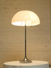 Load image into Gallery viewer, A modern table lamp with a glossy chrome base and stem, featuring a large, dome-shaped off-white shade that glows warm white when lit. The lamp is shown turned on, sitting on a round white pedestal against a textured, light beige brick wall.