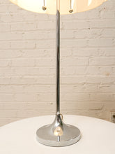 Load image into Gallery viewer, A modern table lamp with a polished chrome base and stem, a warm white fabric drum shade, and pull-chain switches, set on a white round table against a white brick wall.