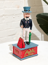 Load image into Gallery viewer, Vintage Uncle Sam figural piggy bank wearing a top hat, dark jacket, bow tie, striped trousers, and standing on a red and blue base