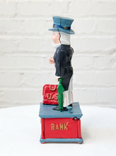 Load image into Gallery viewer, A vintage-style bank toy figurine depicting a postman standing on a red and blue box with the word 'BANK' on the front. The postman wears a blue hat and coat, holds a green umbrella, and stands next to a red mailbox.